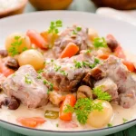 Blanquette de veau servie dans une assiette blanche avec sauce crémeuse, carottes, oignons grelots et champignons